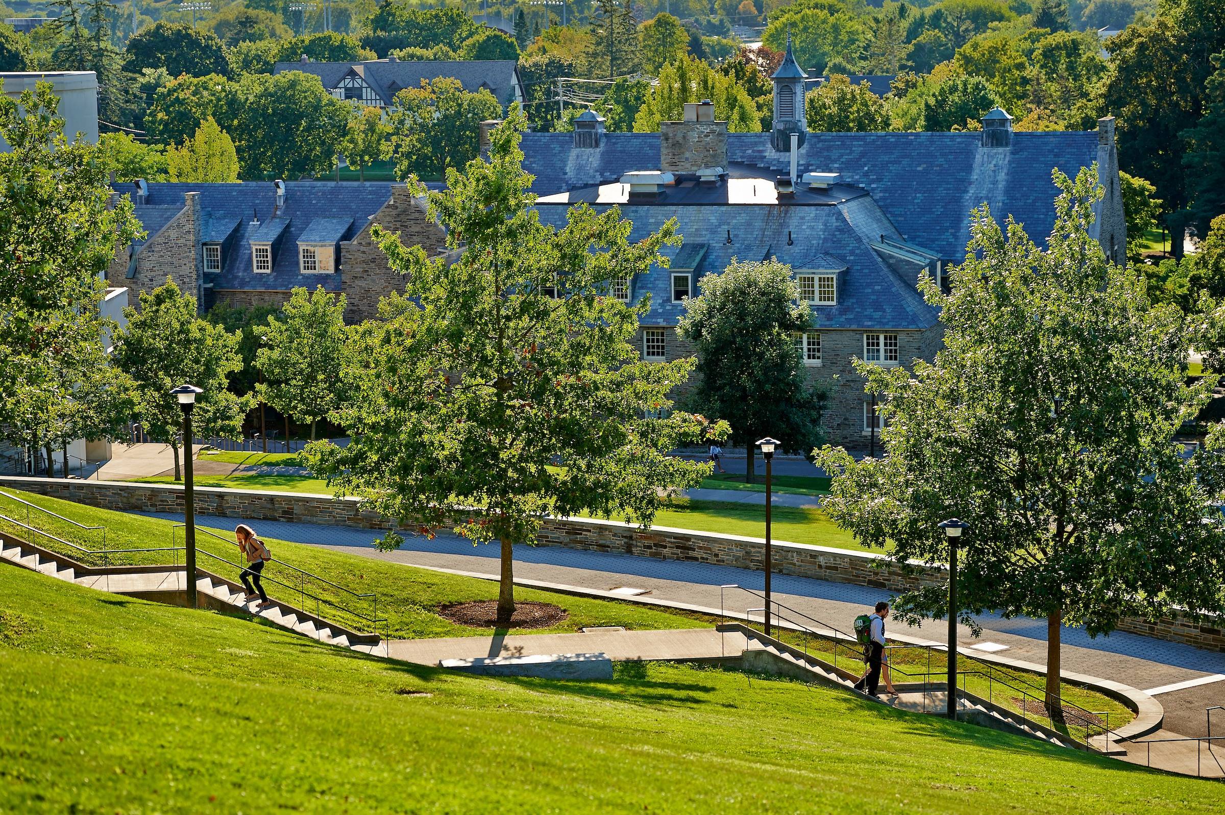 View of Colgate University with lush green lawns and paved walkways. People are seen walking among the trees. Traditional stone buildings with gabled roofs fill the background, bathed in sunlight.
