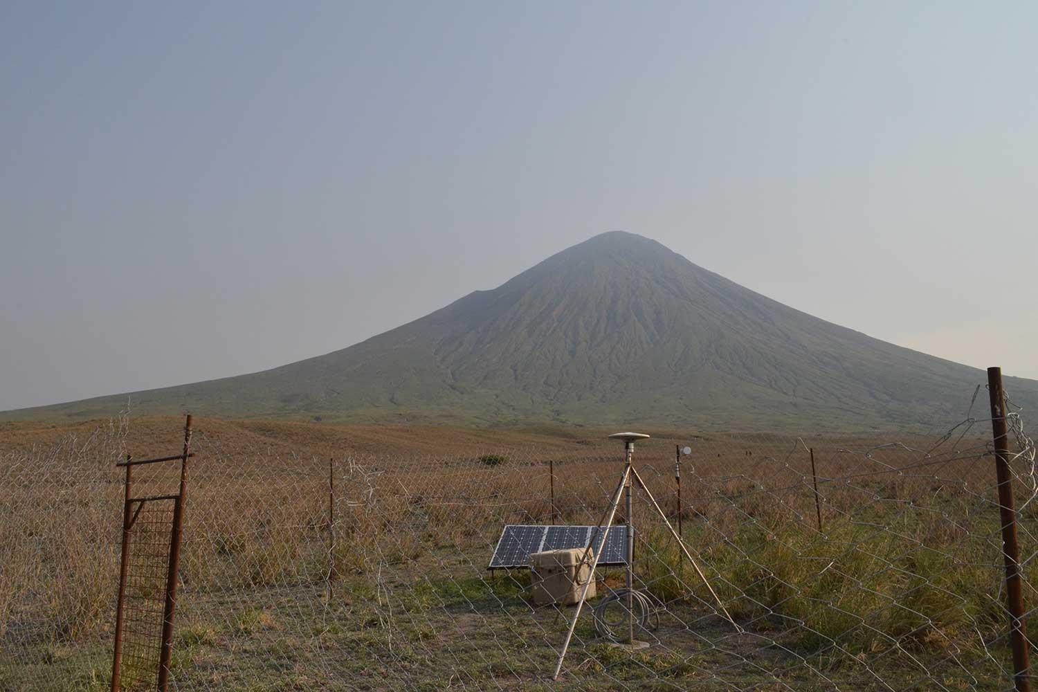 a fenced-in area holding a tripod and other pieces of equipment. Ol Doinyo Lengai is in the distance.